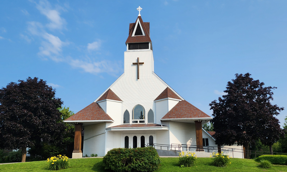 The white wooden exterior of St. Thomas Anglican Church, with its steeple and cross, an arched stained-glass window over the entrance, and trees on either side.