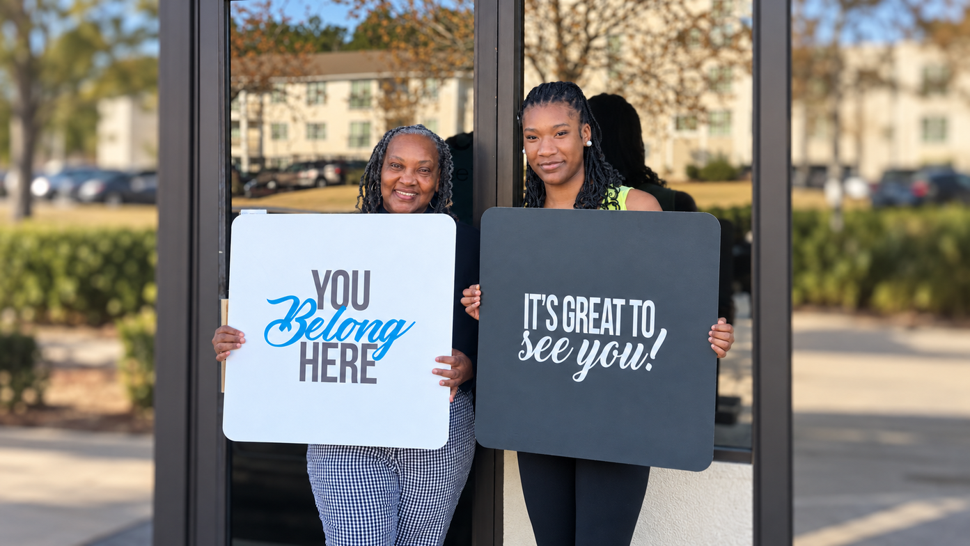 Two smiling welcome team members holding You Belong Here and It&rsquo;s Great to See You signs